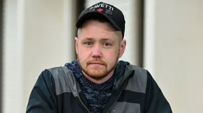 Натан Ньюби, 30-летний мужчина в темной одежде и черной бейсболке, смотрит в камеру. 