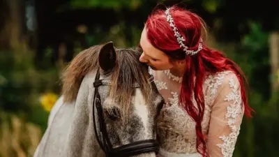 A woman with red hair in a wedding dresses holds her face next to a white-grey horse