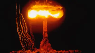A nuclear explosion on 4 June 1953 at the Nevada Proving Ground in the US. A mushroom cloud filled with orange flames at the top rises above the ground. Thick clouds fill the ground. Several streaks of debris appear to one side of the cloud. The surrounding sky is dark. The test is described Operation Upshot Knothole, CLIMAX Event, when a 61 kiloton (55 kilotonne) device was detonated