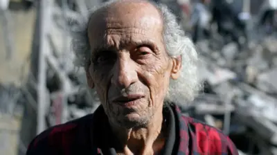 A man, Fouad Hassan, stares off into the distance. Behind him is a twisted pile of concrete and metal rubble. Some of his family used to live where the rubble is now piled high.