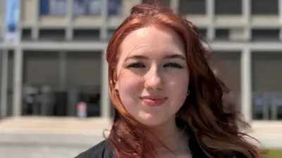 Amelie with shoulder-length red hair in the foreground, facing the camera outdoors, wearing a black short-sleeved shirt and small facial piercings. Behind, a large modern concrete building with rectangular windows and tall vertical columns spans the width of the image. Several flagpoles with light-coloured flags stand near the entrance. A wide paved area leads up to the building, bordered by low greenery. The sky is bright blue with scattered white clouds.
