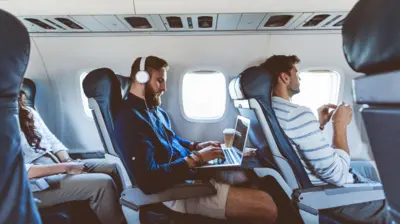 Bearded man in headphones typing on a laptop seated on an aeroplane