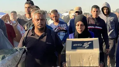 GHF logolu yardım kutuları ve çaresizlik içindeki Gazzeliler aynı karede. Elinde kutu olan bir kadın yanında ve arkasında erkeklerle ilerliyor.