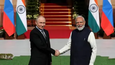 インドとロシアの国旗を背に、明るい日差しの中でダークスーツ姿のプーチン氏と、黒いベストにクリーム色のシャツのモディ氏が、笑顔で握手をしながらカメラを見ている
