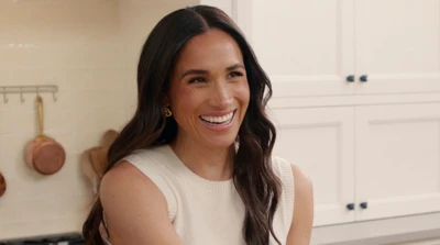 The Duchess of Sussex pictured wearing a white sleeveless jumper smiling a she places a cup on the kitchen counter