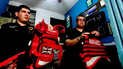 Dos paramédicos, Julio César Vega y Héctor Torres, están sentados en una pequeña habitación. Llevan su equipo sobre las rodillas. Ambos visten uniformes negros y Torres lleva gafas.
