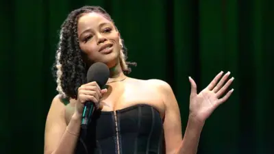 Kyrah Gray performing on stage for the BBC 1Xtra Comedy Gala at the Hackney Empire on 1 April. Kyrah is a black woman in her 20s with brown eyes and dark hair worn loose with a platinum blonde streak. She wears a black corset-style top paired with camo-green trousers. She holds a microphone with her right hand while her left is extended out to her side. The staging behind her is dark with BBC Radio 1Xtra branding in lights.