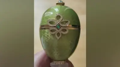 Zbliżenie zielonej ozdoby w kształcie jajka Fabergé ze złotą opaską pośrodku i dekoracyjnym wzorem w kształcie diamentu z małym niebiesko-zielonym kamieniem szlachetnym.