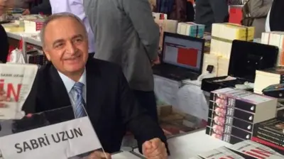 Sabri Uzun kitap fuarında, masada oturuyor, kameraya gülümsüyor