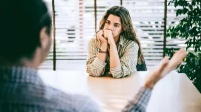 Uma mulher em um escritório está sentada em frente a um homem, que aparece de costas, desfocado ao fundo; ela tem expressão de irritação, e ele gesticula 