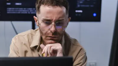 Um engenheiro de software, vestindo uma camisa marrom clara, olha para um monitor de computador à sua frente com uma expressão preocupada. Uma tela maior com código de computador está em uma parede atrás dele.