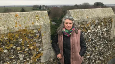 Monique Bonney in front of a stone wall. Behind her is a view of north Kent, including fields, a house and in the distance a town. 