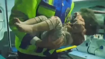 Uma menina pequena é segurada nos braços de um socorrista perto de uma ambulância