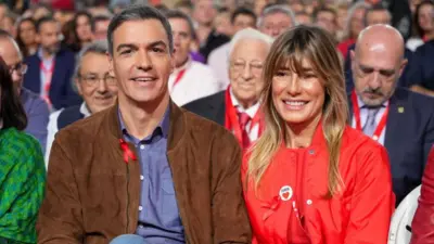 Kırmızı rahat giysi giymiş bir kadınla, yine rahat bir blazer ceket ve mavi gömlek giymiş bir adam yan yana oturuyor. Arkalarındaki koltuklar da dolu