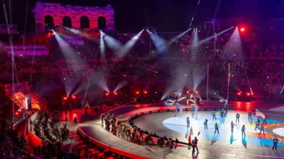 円形闘技場の内部で、観客席が紫色の照明で照らされる中、選手らが入場している。中央は明るく照らされ、米国旗を担ぎ歩く人が見える