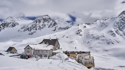 O albergue "Schöne Aussicht Bella Vista". Imagem mostra chalé alpinoslots party paga mesmodia, rodeado por montanhas nevadas sob céu azul. Esquiadores estão no local, com destaque para a cúpula branca e o terraço com vista panorâmica.