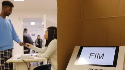 Imagemviver de apostas onlineuma urna eletrônica com duas pessoas, desfocadas, atrás: um eleitor e uma mesária