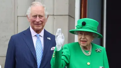 O então príncipe Charles, em um terno azul com listras, ao lado da rainha Elizabeth 2ª, que acena vestindo chapéu e traje verdes