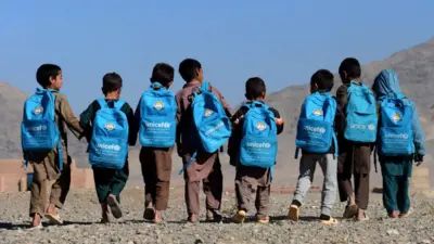 چند کودک که بیگ‌های مکتب با نشانی یونیسف در پشت شان است در حال راه رفتن هستند.