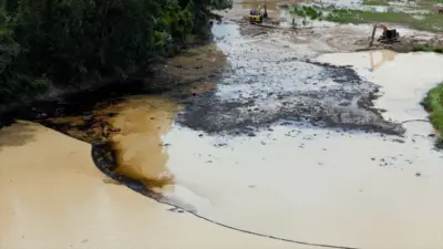 Area of light brown water with black, oily-looking substance along the bank and partially along a containment boom that runs across the water. Dark mud and two diggers can be seen in the background.