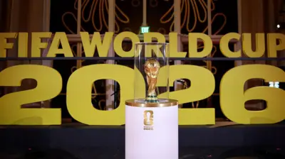 Di World Cup on display on white stand prior to di draw in December in Washington wit Fifa World Cup 2026 in big gold letters for di background.