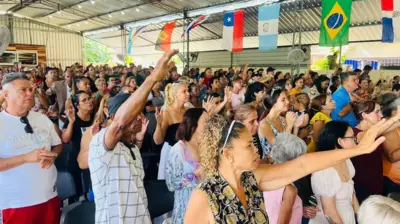Pessoas com braços erguidos em igreja, em ambiente iluminado e com bandeiras de vários países, inclusive o Brasil