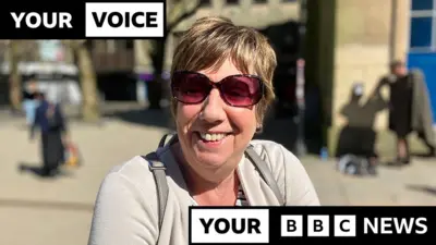 A woman with short brown hair is smiling at the camera, wearing sunglasses, a grey cardigan and a grey backpack