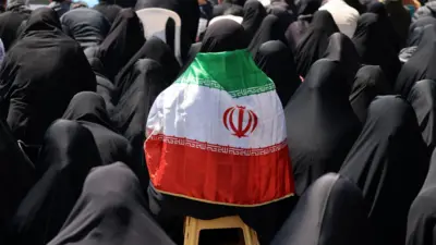 An Iranian woman wears a black hijab and chador and has a national flag draped over her shoulders as government supporters gather at Enghelab Square following the US-Iran ceasefire announcement, in Tehran, Iran (8 April 2026)