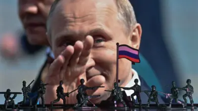 Arka planda, Rusya Devlet Başkanı Vladimir Putin'in koyu mavi ceket ve beyaz gömlek giymiş halde elini salladığı yakın çekim bir fotoğraf görülüyor. Ön planda ise, yatay beyaz, mavi ve kırmızı çizgili minyatür bir Rus bayrağı ve bayrak direğinin etrafında 10 koyu yeşil oyuncak asker yer alıyor.