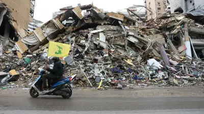 یک موتورسوار با پرچم حزب‌الله لبنان از کنار ویرانه‌های ساختمان‌ها می‌گذرد