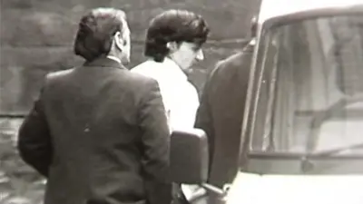 A black and white still from TV footage showing Peter Sullivan, who has thick black hair and is wearing a white shirt, being led to a police van in handcuffs.