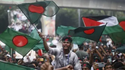A young person wearing a headband displaying the Bangladeshi flag waves a large Bangladeshi flag and shouts slogans. The flag is dark green with a red circle in the centre. Dozens of protesters surround him, most wearing identical headbands, during the Anti-Discrimination Student Movement rally to mark one month since the ousting of Bangladesh's former Prime Minister Sheikh Hasina, in Dhaka, Bangladesh, on 5 September 2024.