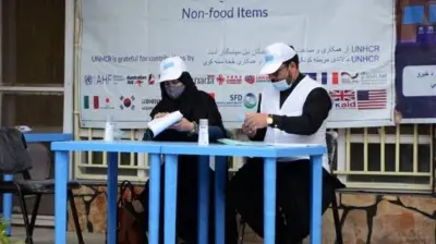 تصویر دو کارمند ملل متحد در یکی از مراکز توزیع کمک‌های نقدی. این کارمندان پشت میز آبی نشته و کلاهی با نشانی ملل متحد به سر دارند. در عقب شان یک بنر ملل متحد دیده می‌شود. 