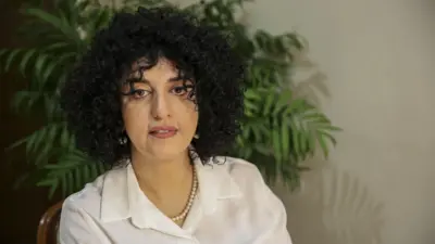 白いブラウスに真珠のネックレスとイヤリングを着けた、黒い巻き毛の女性の写真。背後には観葉植物が置いてある