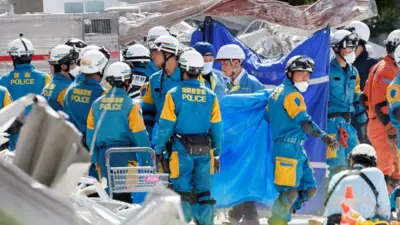 がれきの散乱する中、さまざまな熊本県警などさまざまな所属の隊服を着た人たちたヘルメットやマスクをつけて立っている