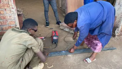 Françoise Umuhoza, wambaye itabuliya y'ubururu n'indorerwamo zijimye, agaragara ateranya ibyuma mu kazi ke akora ko gusudira.