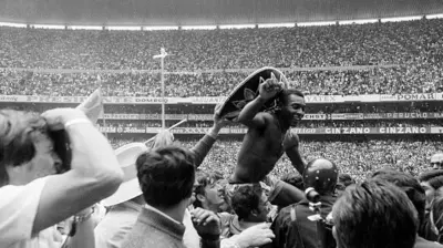 A foto em preto e branco mostra Pelé sendo carregado nos ombros por uma multidão dentro de um estádio lotado. Sem camisa e usando um grande sombrero, ele ergue o dedo indicador enquanto sorri para o público, cercado por torcedores, fotógrafos e seguranças. Ao fundo, as arquibancadas completamente cheias e os painéis de publicidade reforçam o clima de celebração após uma partida.