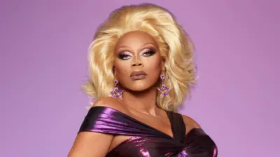 RuPaul in a promotional shoot for the sixth series of RuPaul's Drag Race UK. RuPaul wears a metallic purple off-the-shoulder dress, paired with a blonde wig and purple star-shaped earrings. She pouts and poses against a purple backdrop. 