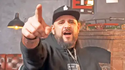 El Temach usa um capuz preto, um boné preto e branco e tem barba. Ele está apontando e gritando para a câmera.