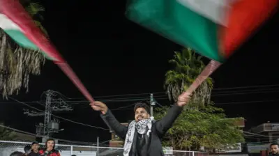 Smiling Palestinian man wearing traditional scarf with arms extended waves two large Palestinian flags in celebration as several men look on