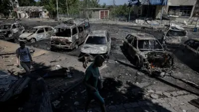 晴天の往来で、路肩に駐車してあった車両7台が焼き尽くされ、路上に粉じんやがれきが散乱する中、住民が歩いている