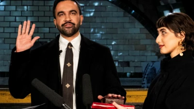 Zohran Mamdani elini kaldırmış durumda ve diğer eliyle de Kuran'a el basıyor. Yanında eşi var