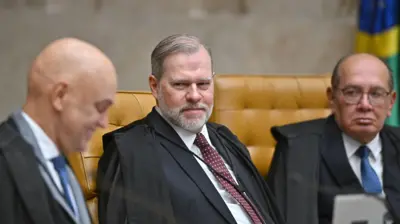 Moraes sorri de perfil ao lado de Toffoli e Gilmar. Os três estão sentados em cadeiras de couro amarelo, no plenario do STF. Eles vestem gravatas, ternos e togas de juiz.