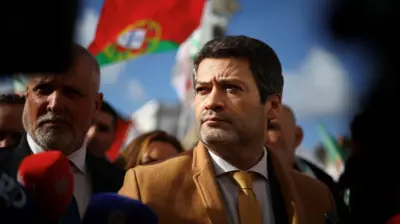 André Ventura olhando para o lado. Há várias pessoas em volta e uma bandeira de Portugal ao fundo