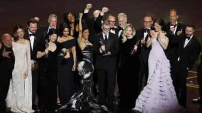 アカデミー賞授賞式のステージ上に集まった15人ほどの人々。女性は白や黒、ピンクのロングドレスを着ている。男性はタキシードにボウタイを着けている。何人かは右手のこぶしを挙げてガッツポーズをしている。