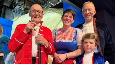 An older man in a red jacket with black rimmed glasses holds a medal and a cane, next to him a woman, a man and a child stand in pantomime costume.