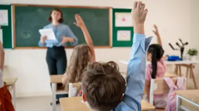 Bir sınıfta ilkokul öğrencisi bir erkek çocuk iki kız çocuk el kaldırıyor. Arkada bir kadın öğretmen elinde kağıtla konuşuyor.