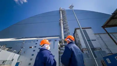 Двое работников ЧАЭС в оранжевых шлемах стоят у купола, защищающего саркофаг