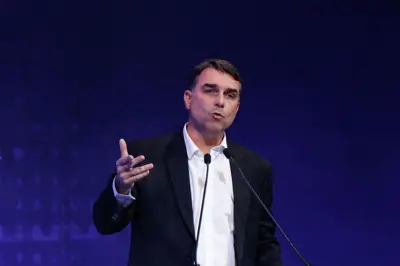 Flávio Bolsonaro aparece de blazer escuro e camisa branca, durante discurso, com um fundo azul