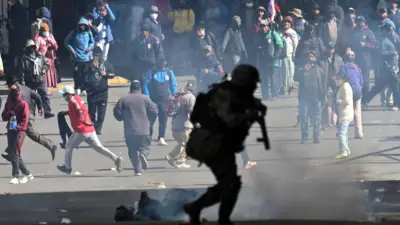Um policial corre em frente a manifestantes durante uma marcha que pede a renúncia do presidente boliviano Rodrigo Paz, em meio ao agravamento da crise econômica e de combustíveis no país, devido à escassez de dólares americanos e à queda na produção interna de energia, em La Paz, Bolívia, 25 de maio de 2026